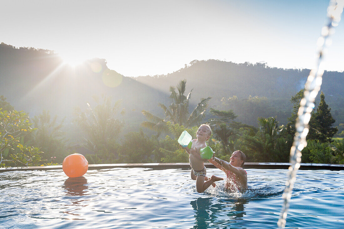 Mother and son playing in a swimming pool, mother throwing boy in the air, splashing water, water ball, sunset, infinity pool, overflow pool, boy 3 years old, palm trees, evening sun, playing, mountains, Balinese holiday resort, family travel in Asia, par