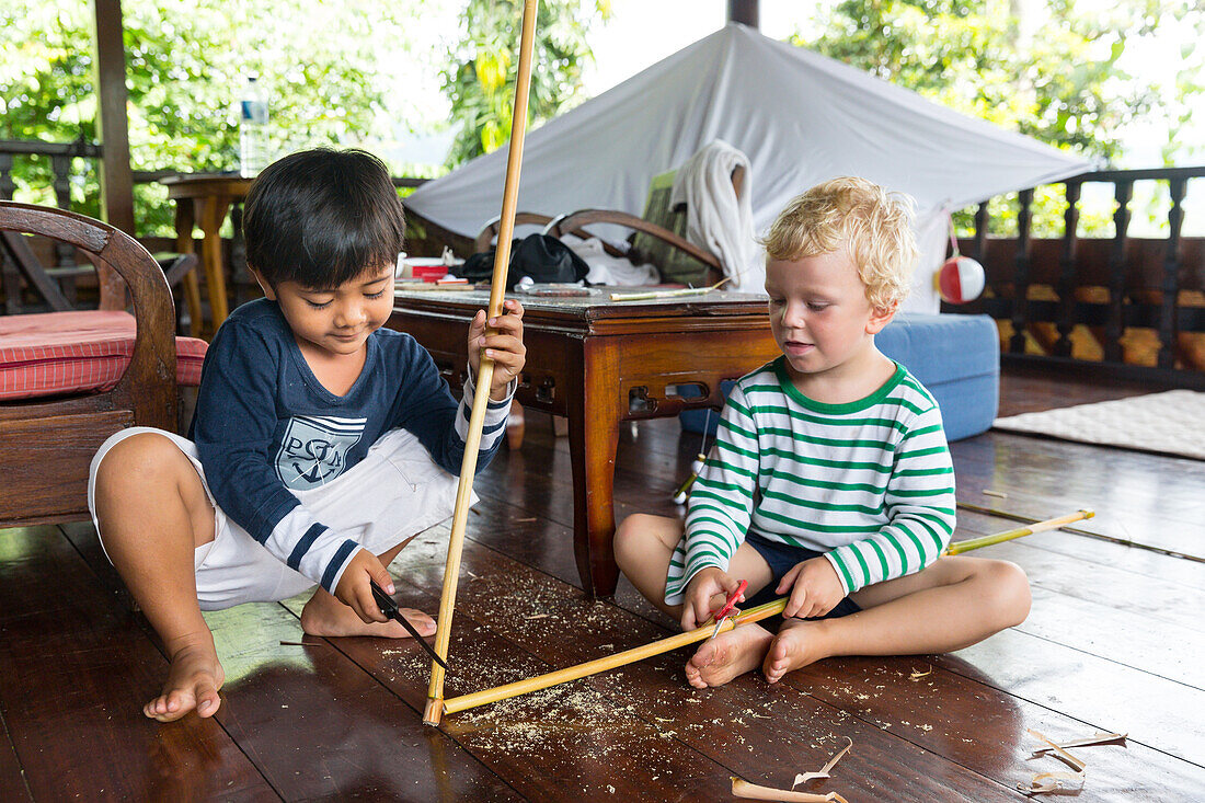Balinese boy playing with foreign boy, German, 3 years old, blond,  knife, whittle, carving a wood stick, wooden terrace, Balinese holiday resort, hotel, family travel in Asia, parental leave, German, European, MR, Sidemen, Bali, Indonesia