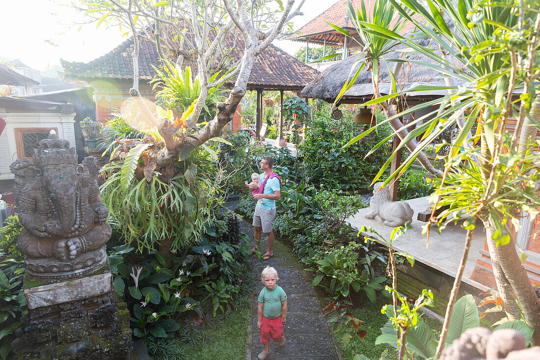 Balinese holiday resort in Ubud, father with baby on arm, 5 months, son,boy, 3 years, blond, tropical garden, sculpture Ganesha, elefant, typical Balinese architecture, hotel, evening sun, family travel in Asia, parental leave, German, European, MR, Ubud,