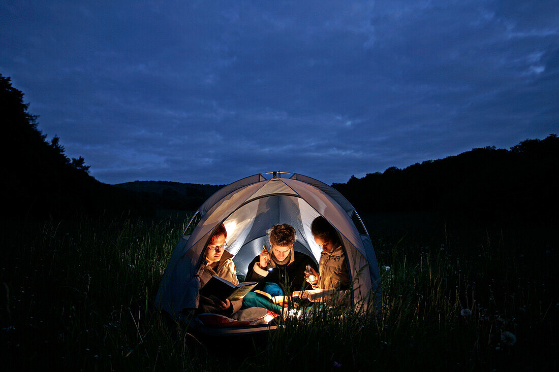 Drei junge Menschen sitzen gemeinsam in einem kleinen Zelt und lesen Bücher im Licht von Taschenlampen, Odershausen, Bad Wildungen, Nordhessen, Hessen, Deutschland, Europa