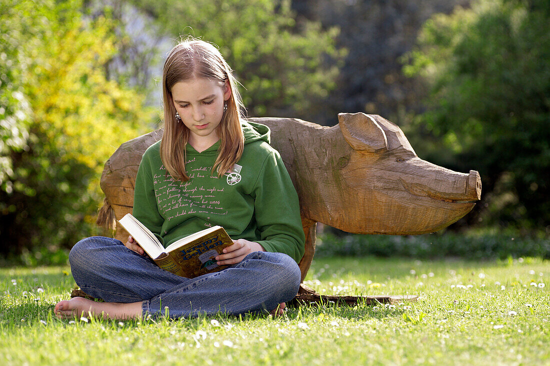 Ein Mädchen liest ein Buch, an ein Holzschwein im Garten gelehnt, Odershausen, Bad Wildungen, Nordhessen, Hessen, Deutschland, Europa
