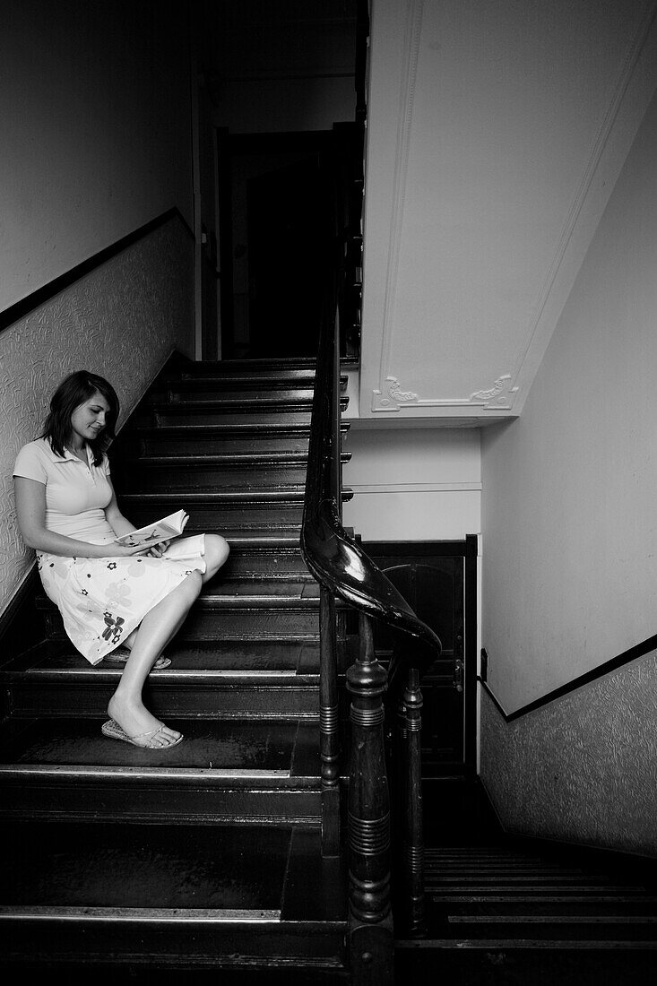 Eine junge Frau sitzt in einem Treppenhaus eines Gebäudes aus der Gründerzeit und liest ein Buch, Berlin, Deutschland, Europa (Schwarzweißaufnahme)