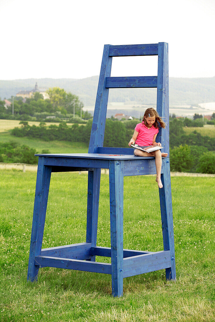 A young girl sitting on an oversized blue chair in a field in front of Schloss Friedrichstein Palace, reading a book, Bad Wildungen, Hesse, Germany, Europe