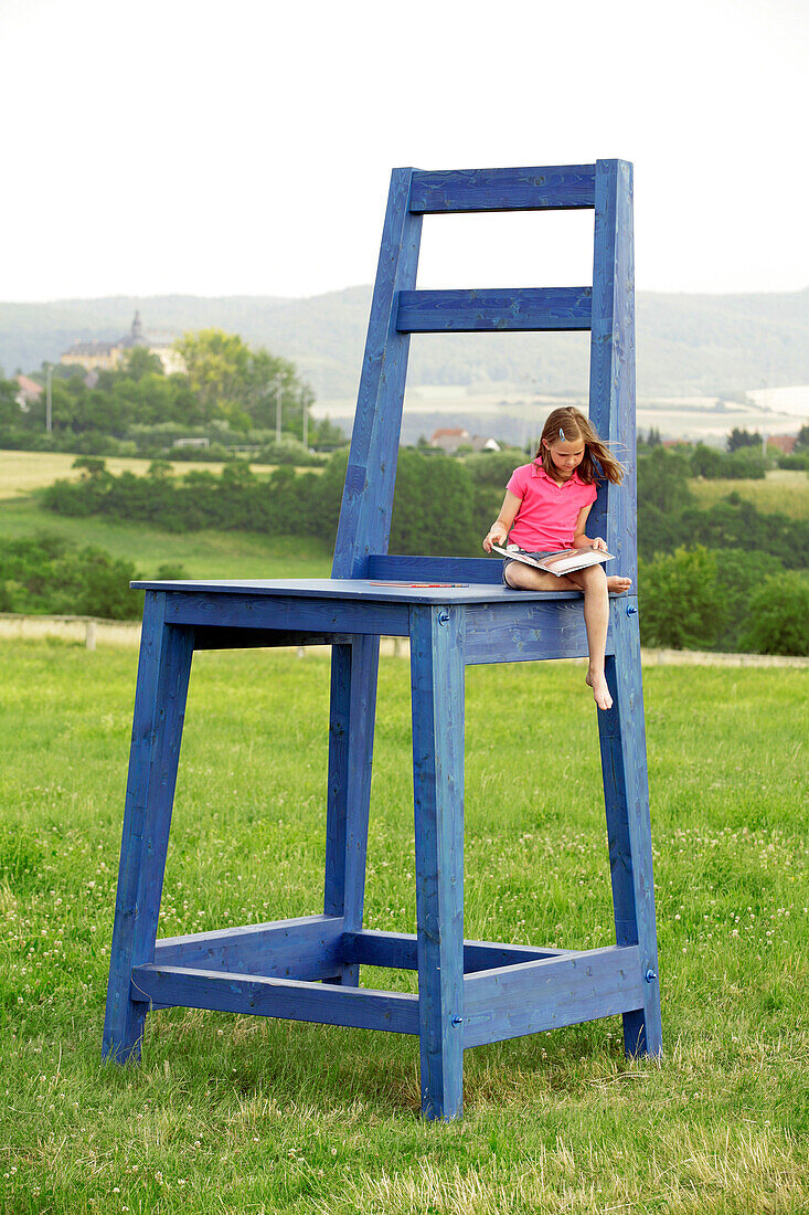 Ein junges Mädchen sitzt auf einem überdimensionalen blauen Stuhl auf einer Wiese oberhalb von Schloss Friedrichstein und liest ein Buch, Bad Wildungen, Nordhessen, Hessen, Deutschland, Europa