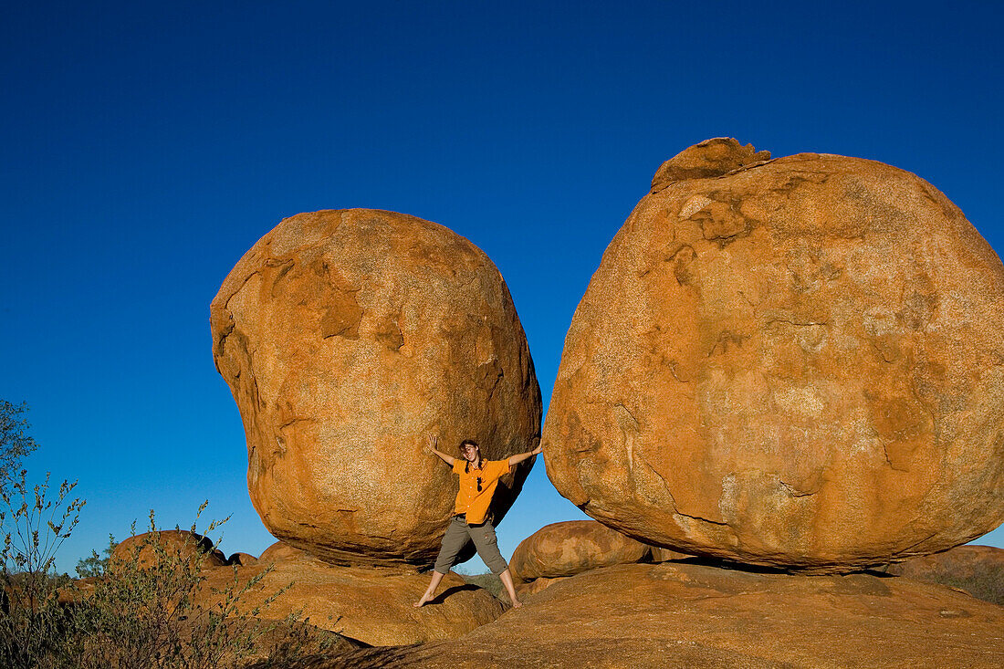 Woman standing inbetween two of the round red rocks called Devils Marbles and pretending to hold them, Devils Marbles Conservation Reserve, near Wauchope, Northern Territory, Australia