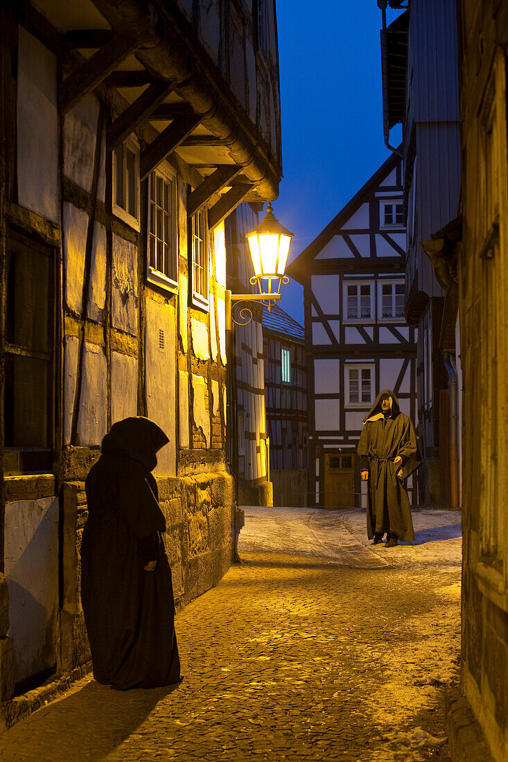 Two people wearing traditional coats meet under a street lamp in a small alley called Paradiesgasse with half-timbered houses at dusk, Bad Wildungen, Hesse, Germany, Europe