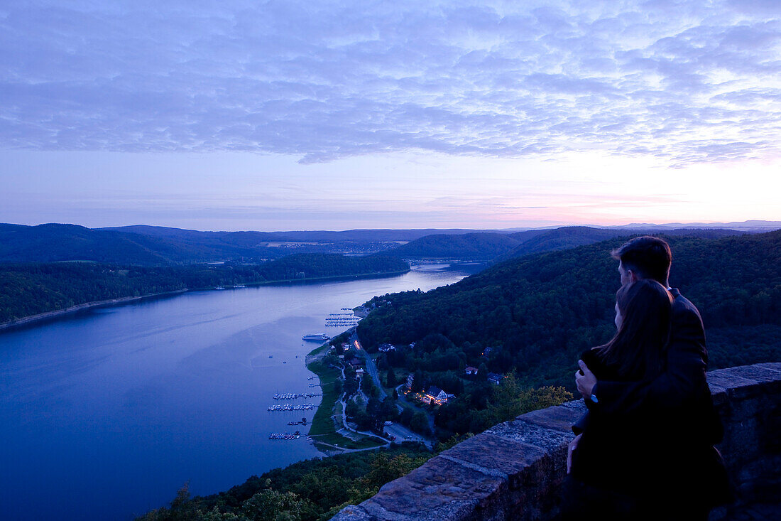 Ein paar steht auf Schloss Waldeck an der Mauer der Aussichtsterasse und schaut auf den Edersee im Nationalpark Kellerwald-Edersee bei Dämmerung, Nordhessen, Hessen, Deutschland, Europa