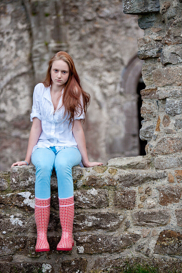 Red haired teenage girl wearing wellington boots with red dots sitting on a stone wall, Clonmacnoise, County Offaly, Ireland, Europe