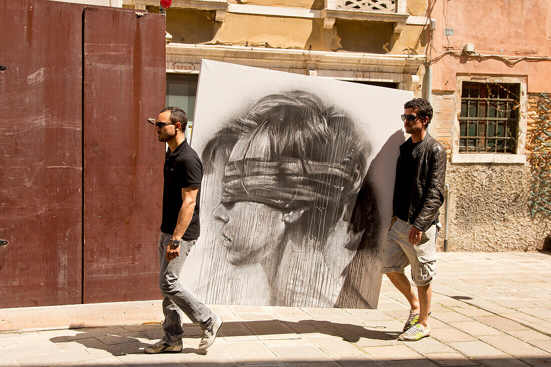 Two young men carrying a huge drawing featuring a woman's head with blindfold along a pathway near Campo Sant Stefano, Venice, Veneto, Italy, Europe