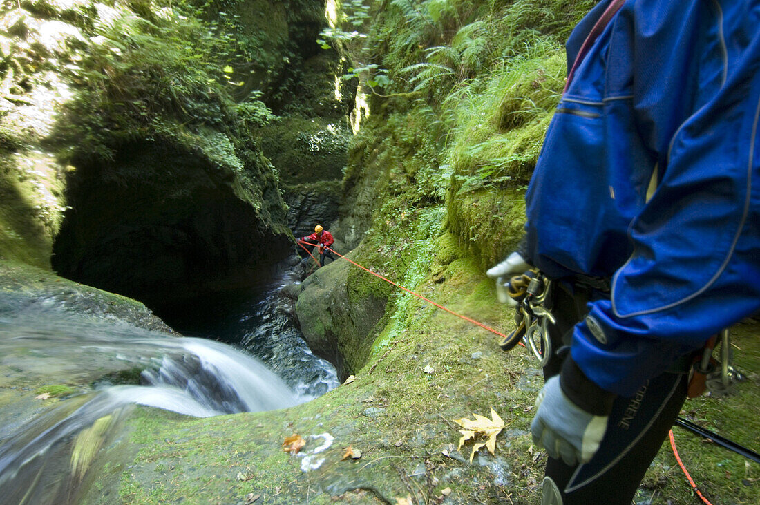 Davis Creek, South of Mount Rainier, WA. Canyoning is a new sport that consists in travelling down river canyons by walking, gliding, climbing, rappelling abseiling, swimming or jumping. Rob Cobb blue, looks at Joe Budgen rappelling abseiling, down past a