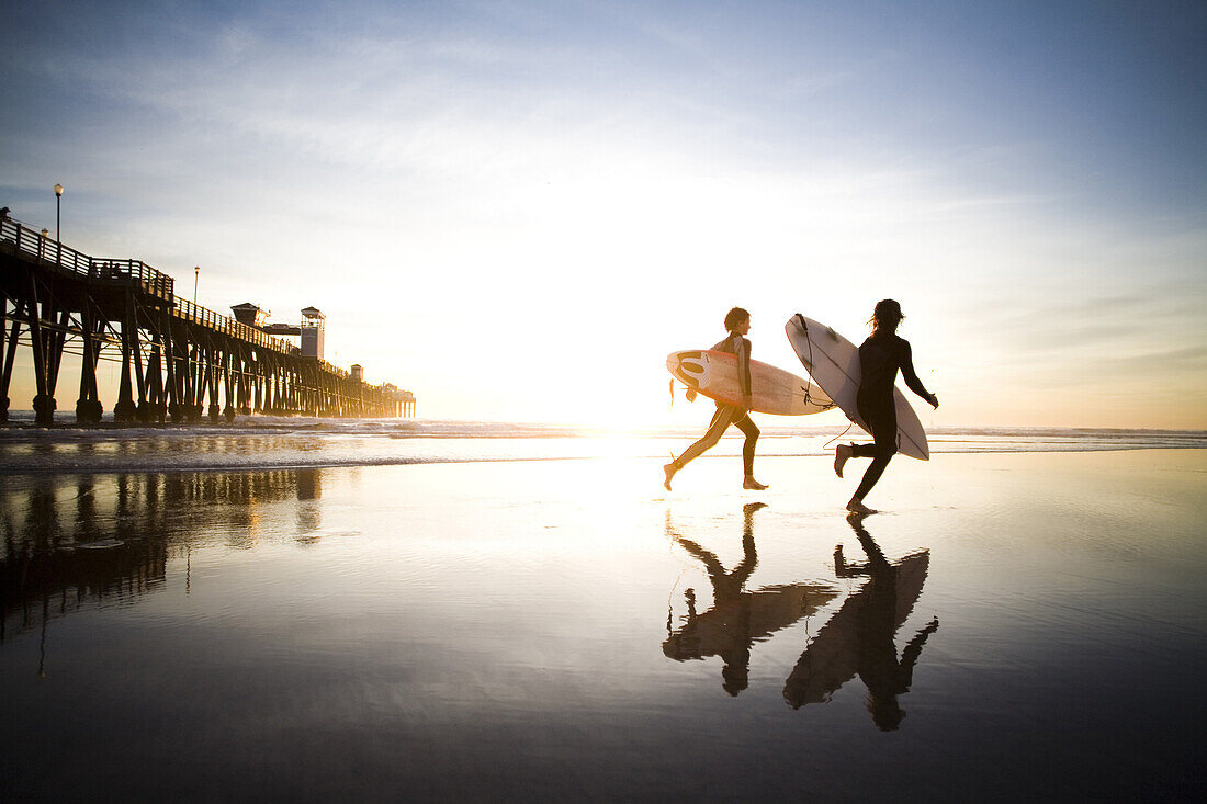 Two surfer run accross the beach at sunset.