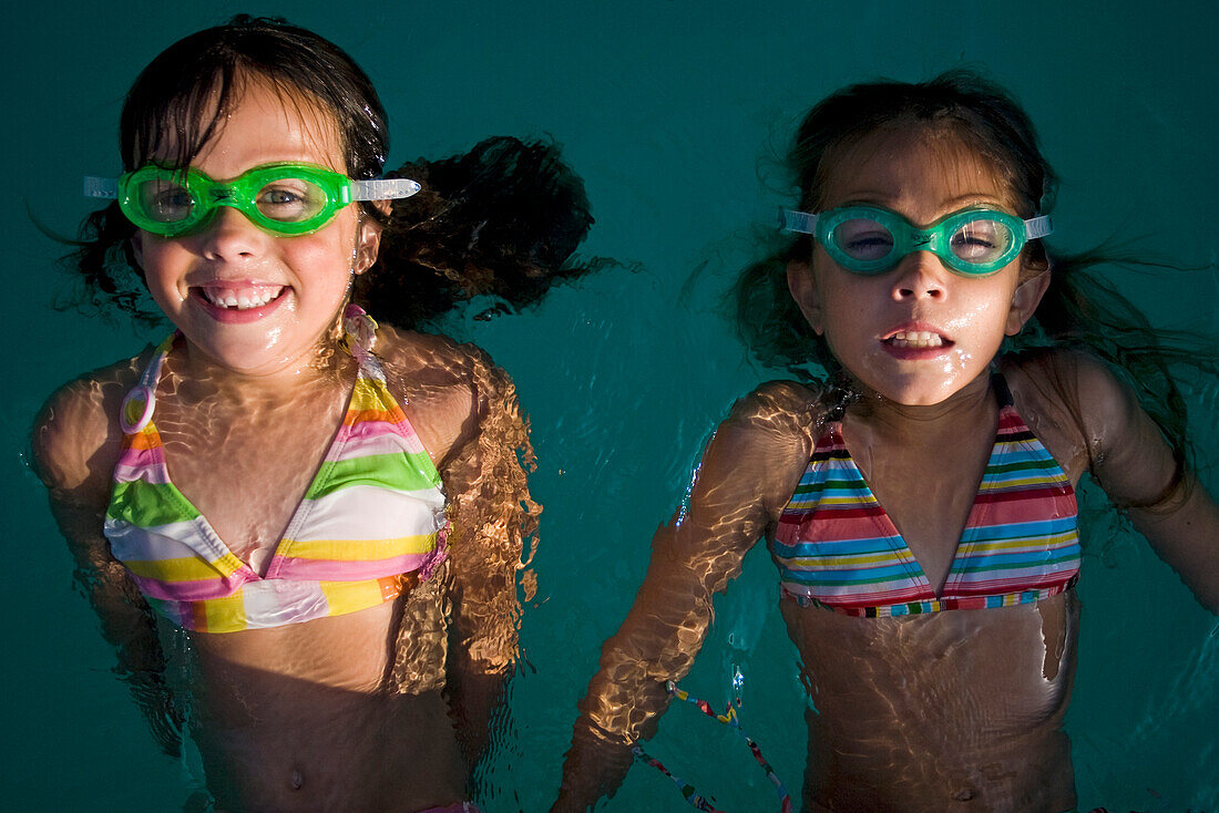 Two girls in goggles float in the water at a pool party.