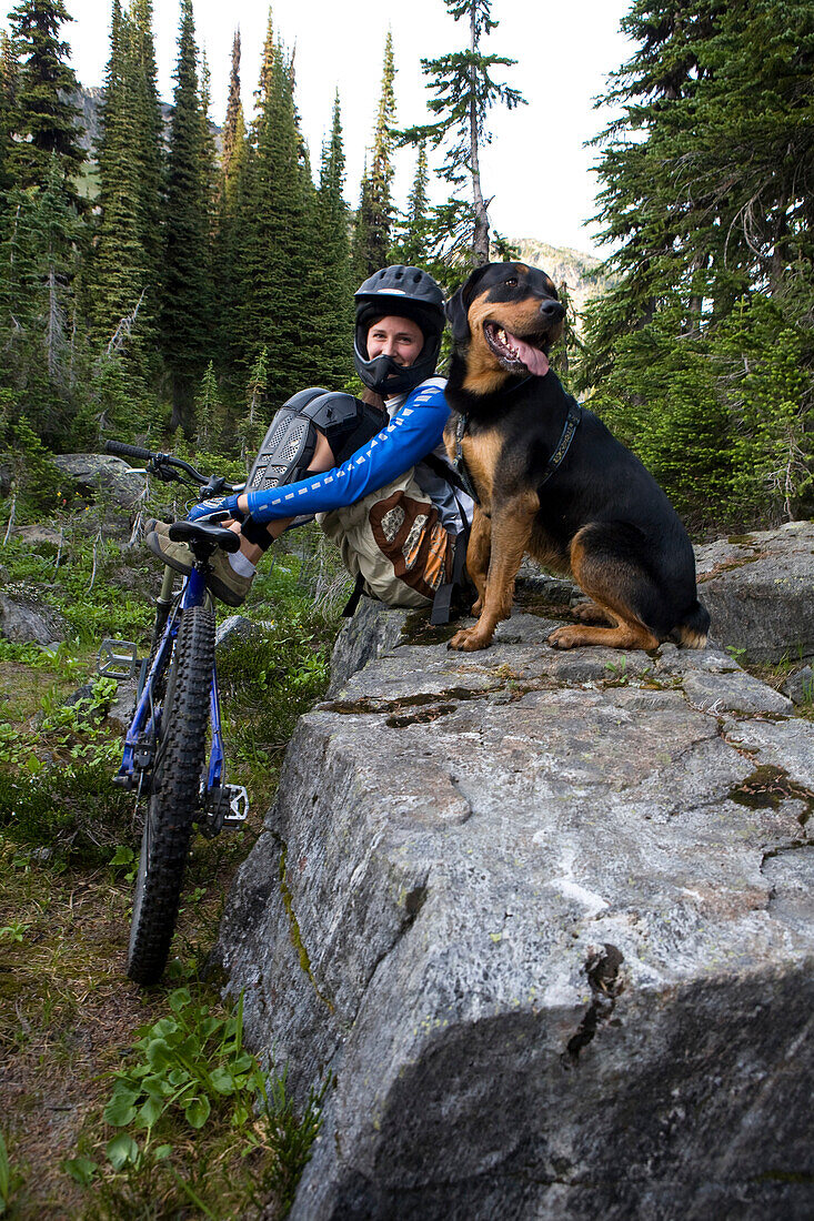 Sol Mountain, BC - Kate Watters and her dog, Babushka, rest as they mountain bike across alpine meadow while flowers in bloom at Sol Mountain Touring's backcountry lodge in the Southern Monashee range of the Columbia Mountains of South Central British Col