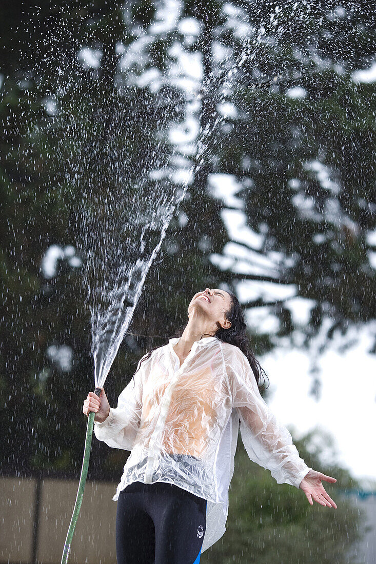A woman squirts water from a hose into the air as she stands under the falling water, San Diego, California. model release code: gibbs_carolyn.jpg