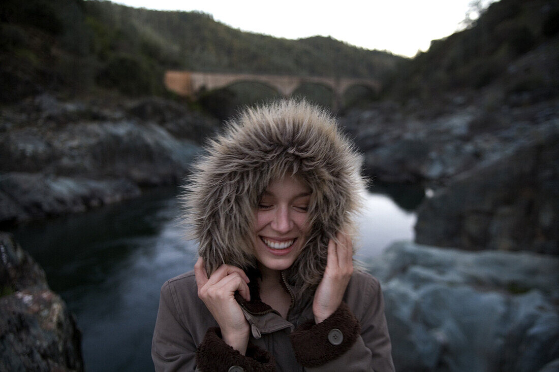 A woman smiles holding the fur of the hood of her jacket in front of a river, Auburn, California.
