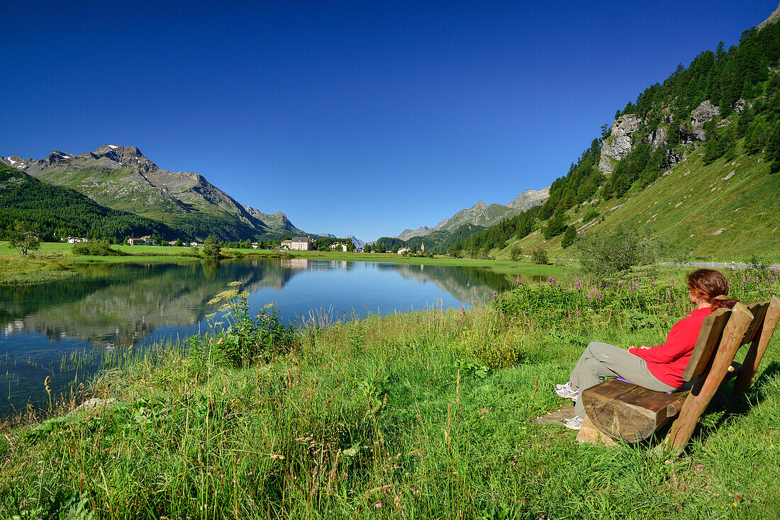 Woman sitting on a bench with view over Inn river to Sils-Baselgia, Sils, Upper Engadin, Engadin, Canton of Graubuenden, Switzerland
