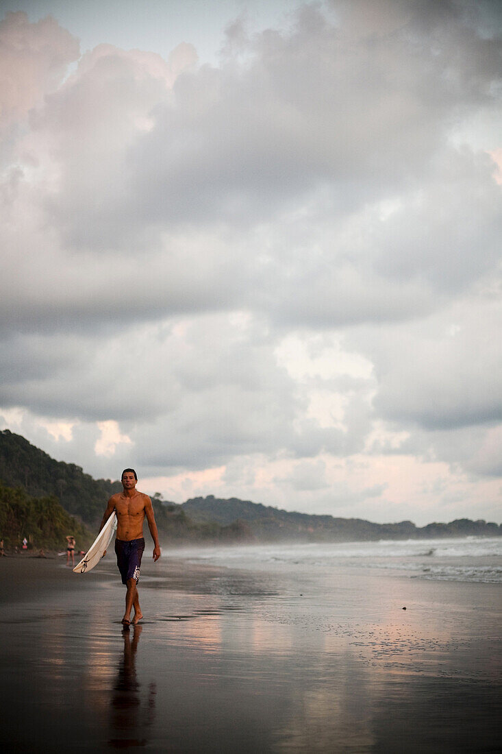 DOMINICAL, COSTA RICA. A man walks down the beach with his surfboard at dusk beneath a cloudy sky.