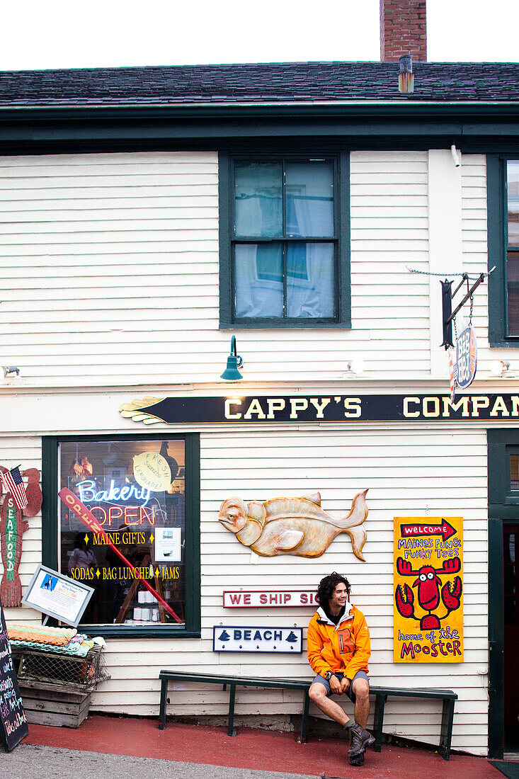 MAINE, USA. A man wearing an orange jacket sits in front of a shop on a small street.