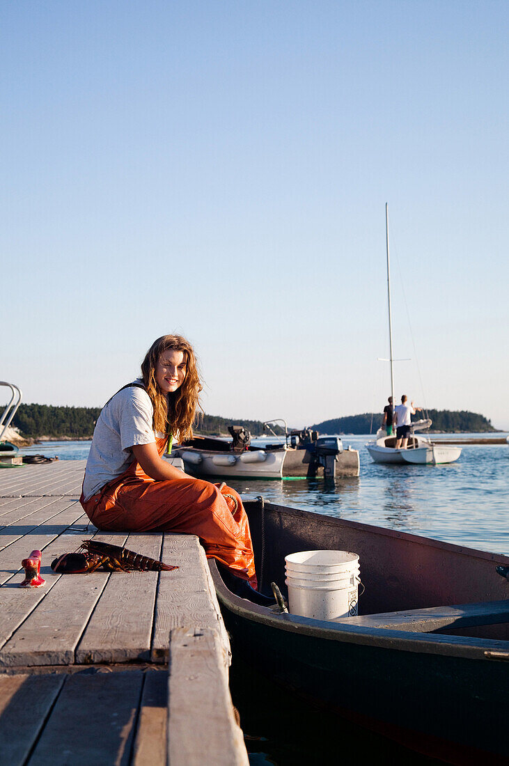 SEBASCO, MAINE, USA. A portrait of a young woman sitting on the edge of a dock with her feet in the boat.