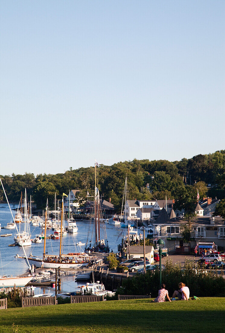 CAMDEN, MAINE, USA. A couple sits on a grassy hilltop overlooking the harbor below.