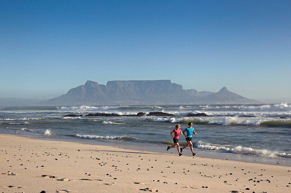 Katrin Schneider and Susann Scheller running along Bloubergstrand beach in Cape Town with a perfect view of Table Mountain in the back. Cape Town, South Africa.