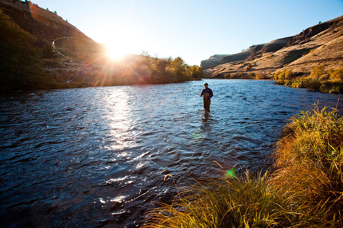 A fly fisherman stands in waist deep water on the Deschutes River with mountains in the distance on a sunny evening outside Bend, Oregon.