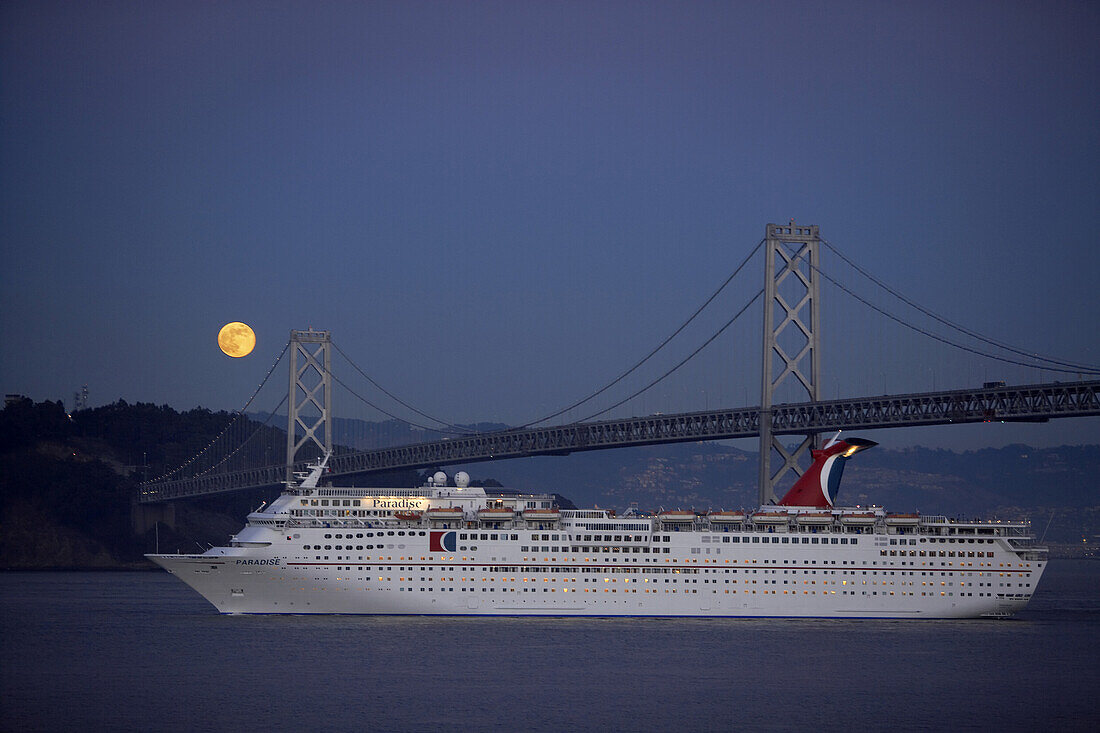 A cruise ship passing under the Bay Bridge in San Fransico with a full moon lighting the night sky.
