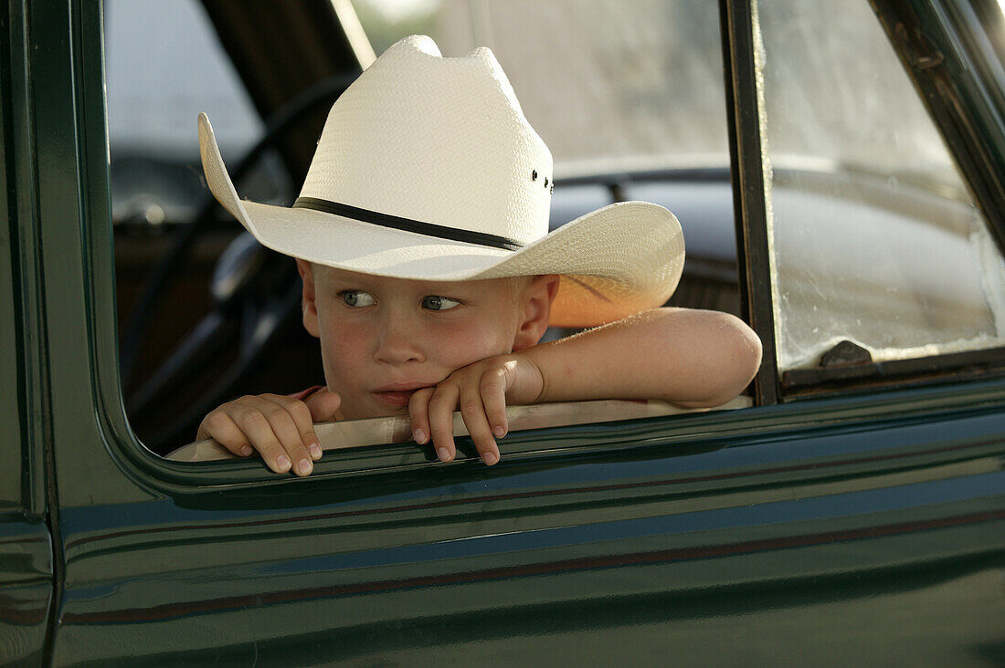 A little boy wearing a cowboy hat leans on the window frame of an old green pick-up truck.