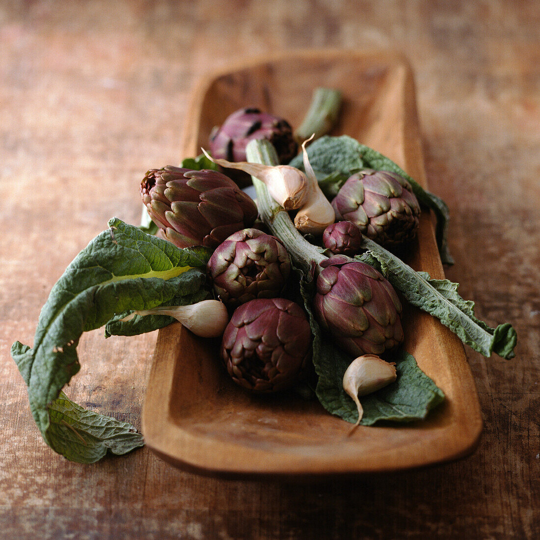 Purple artichokes with leaves and garlic cloves in wooden container, close-up