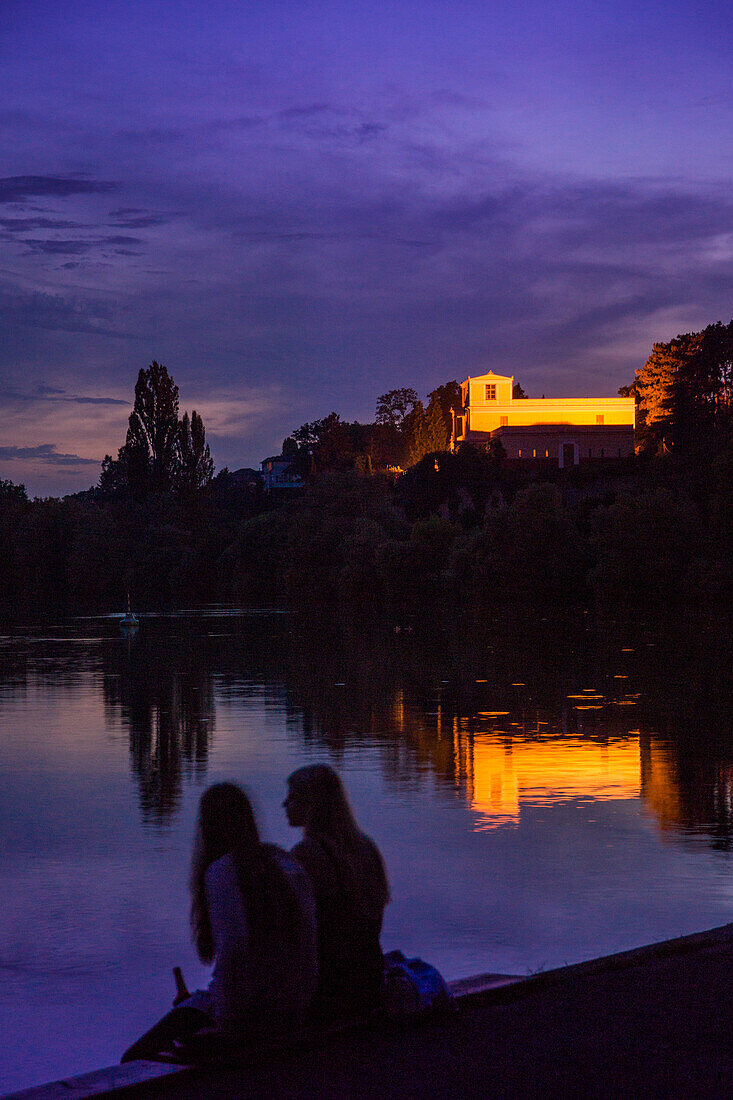 Two young girls sitting on the banks of the river Main at dusk with Pompeiianum Villa near Johannisburg Palace in the background, Aschaffenburg, Franconia, Bavaria, Germany