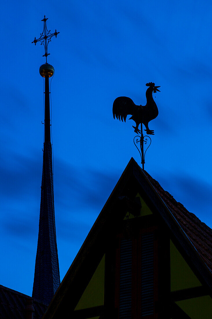Silhouette von Skulptur Hahn auf Dach an Fachwerkhaus mit Spitze von Kirchturm in der Dämmerung, Frickenhausen, nahe Ochsenfurt, Franken, Bayern, Deutschland