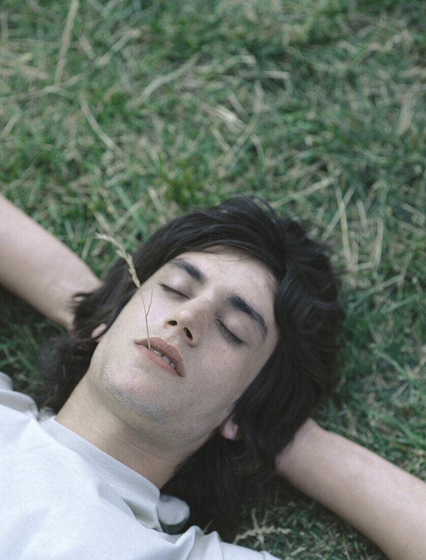 Young man lying on grass with eyes closed and piece of grass in mouth