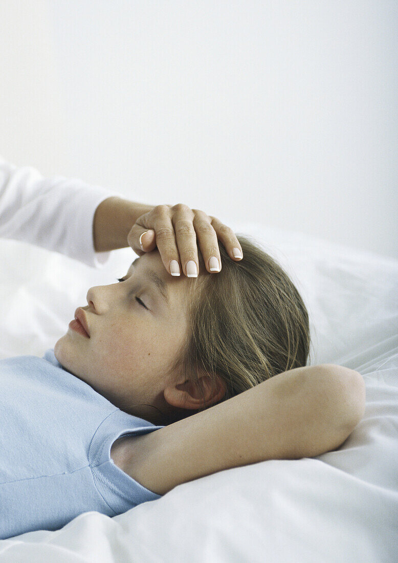 Girl lying in bed with woman's hand on her forehead