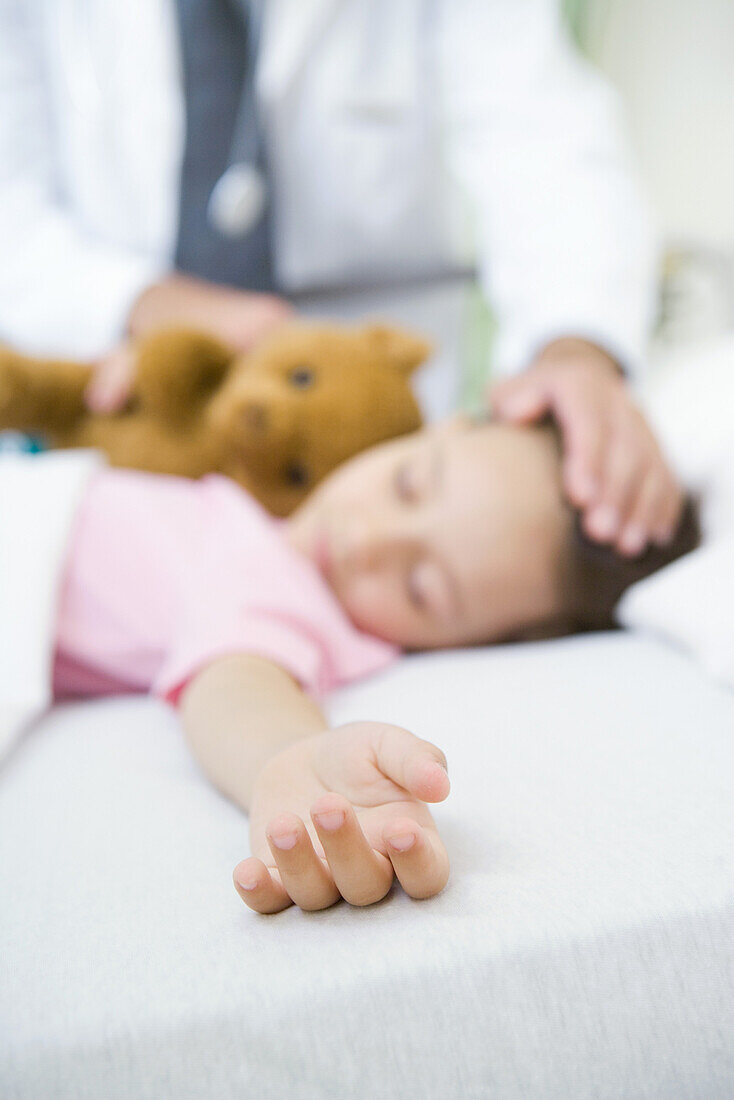 Girl sleeping in hospital bed, doctor's hand on forehead
