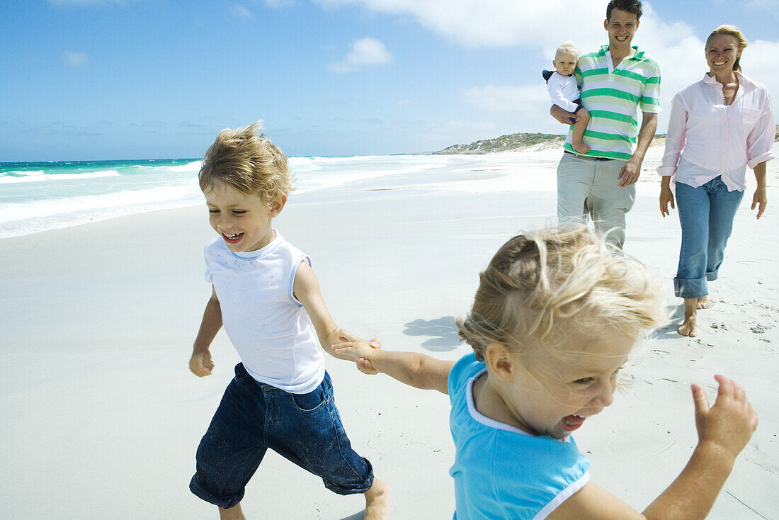 Brother and sister running and laughing on beach in front of parents with baby