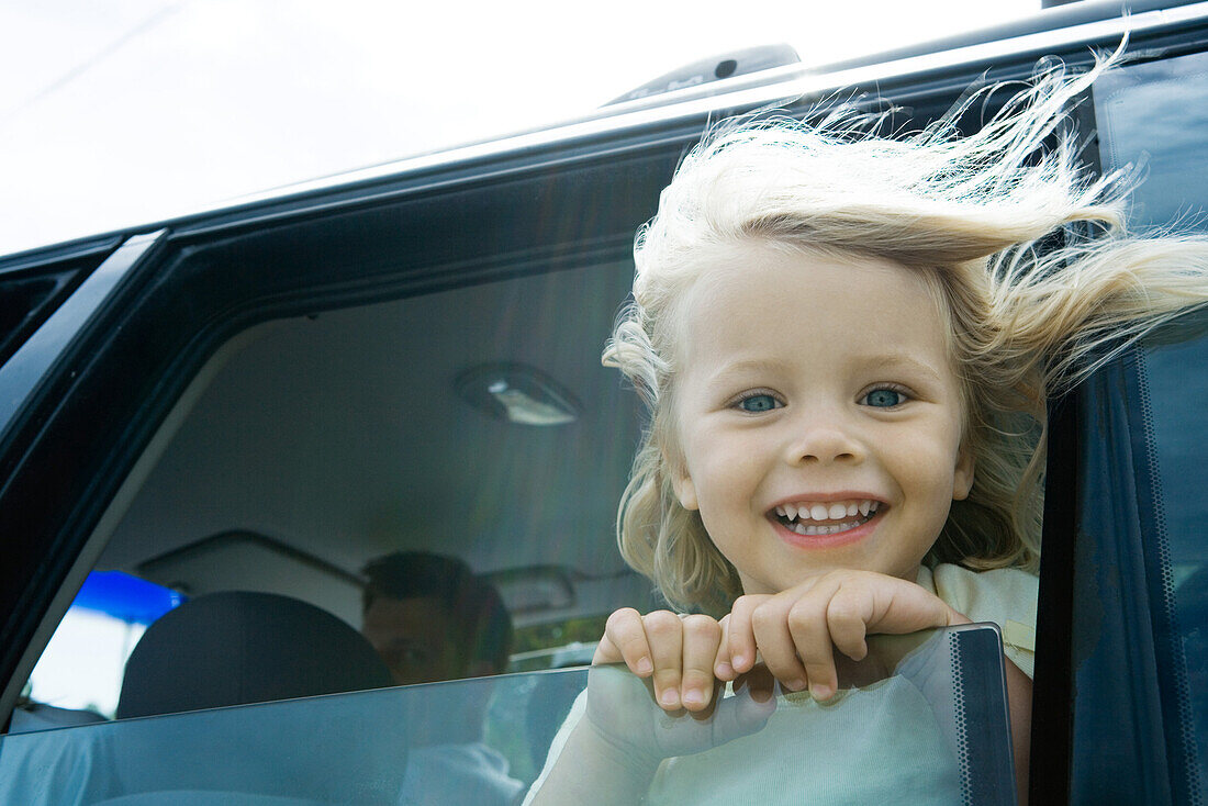 Little girl in car, looking out of window, smiling at camera, hair blowing in wind