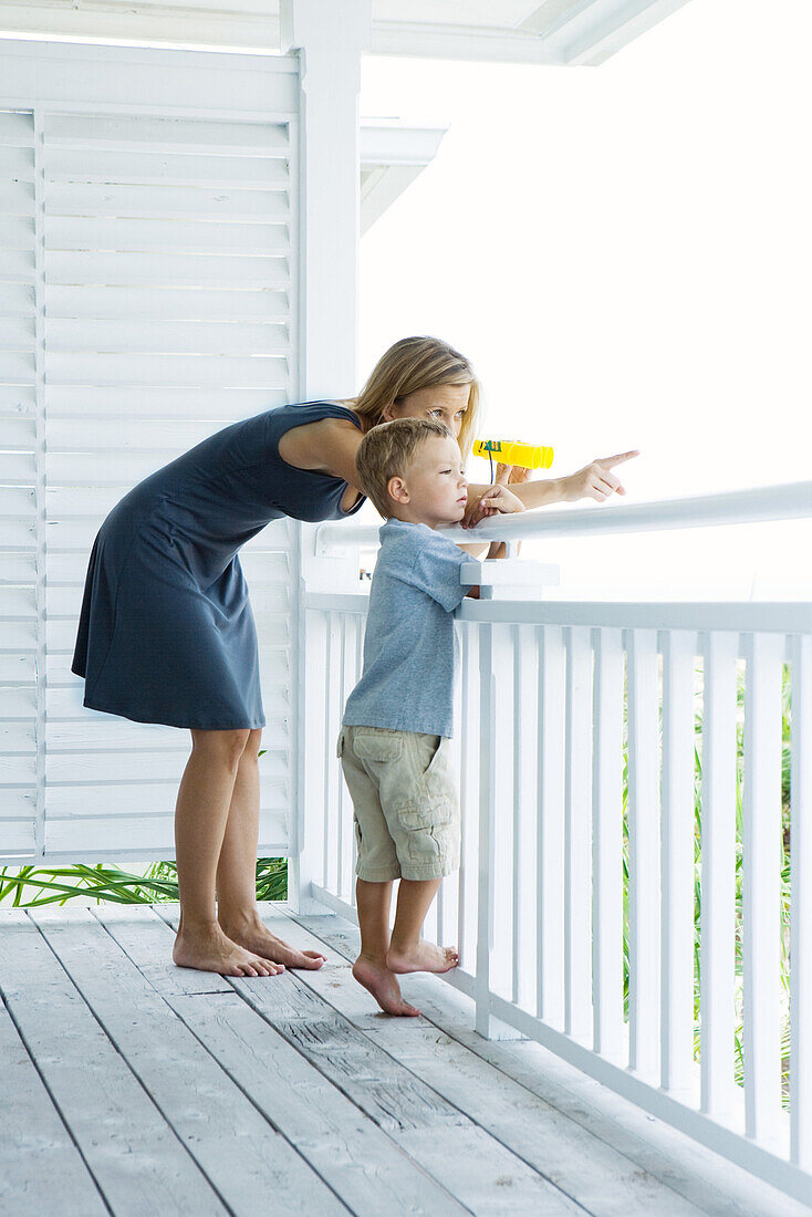 Mother and son standing on porch together, woman holding binoculars and pointing, both looking away