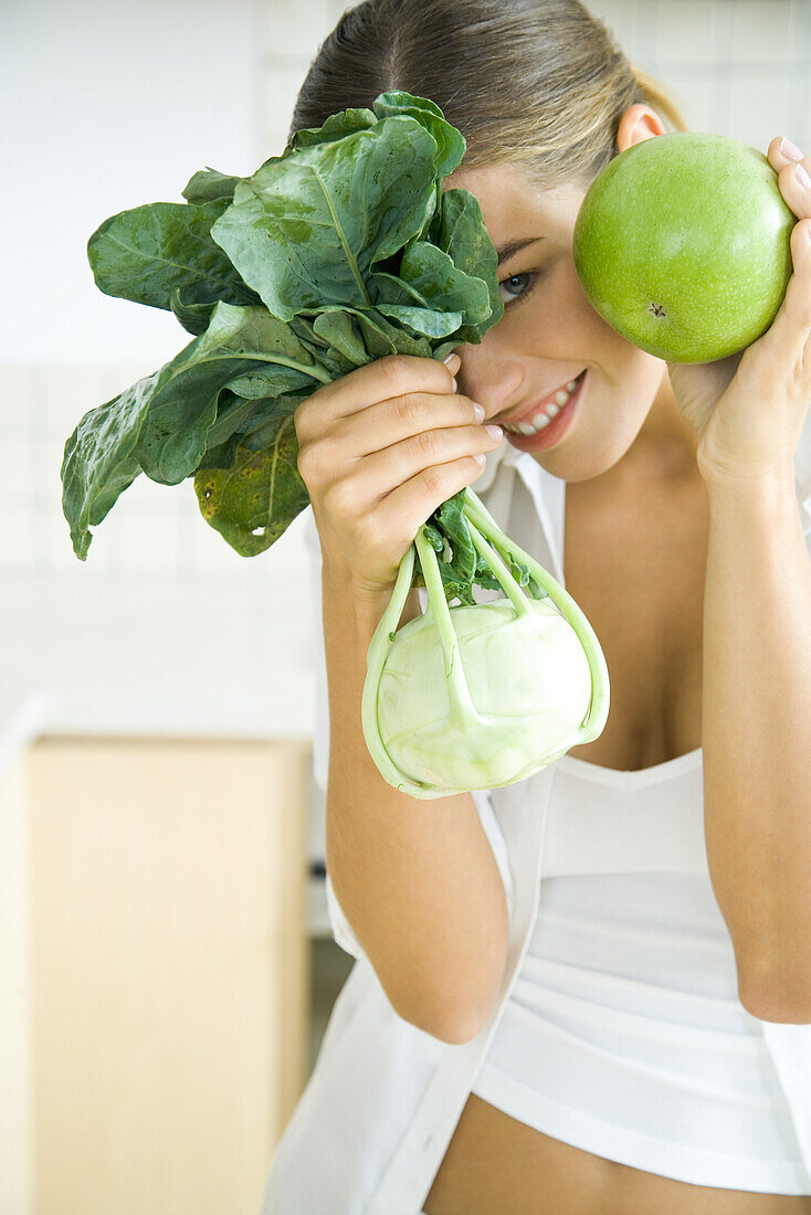 Woman holding up and hiding behind fresh kohlrabi and apple, smiling at camera