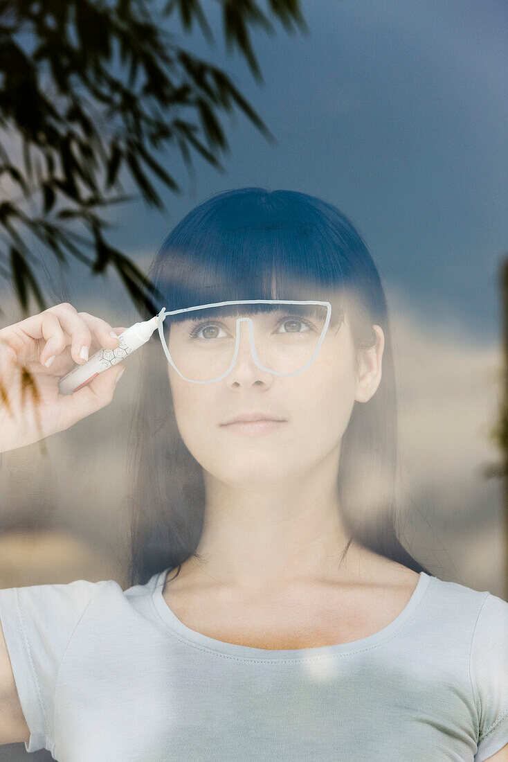 Young woman drawing a pair of glasses on a window pane, looking through window
