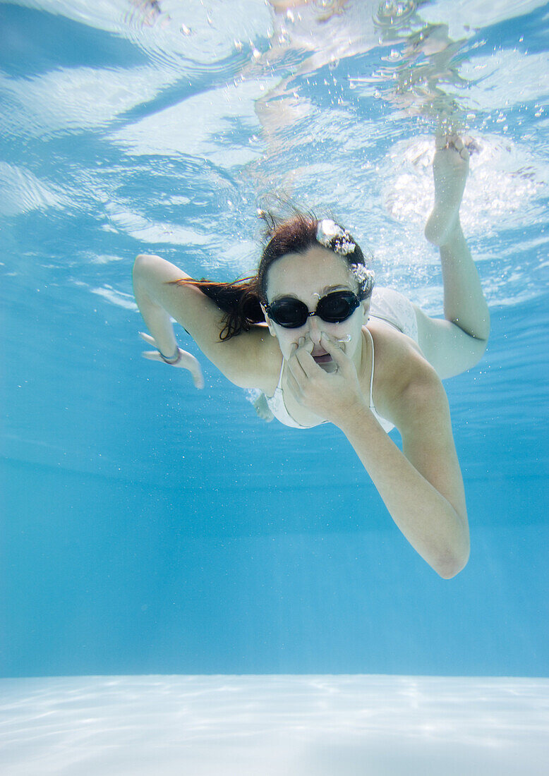 Teenage girl in pool, holding nose and wearing goggles, underwater view