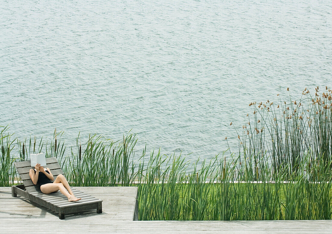 Woman reclining on lounge chair, reading, next to lake