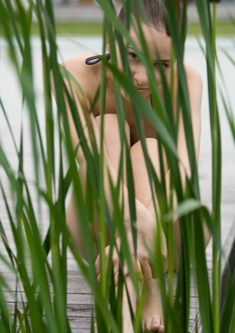 Young woman sitting on deck in swimsuit, reeds in foreground, looking at camera
