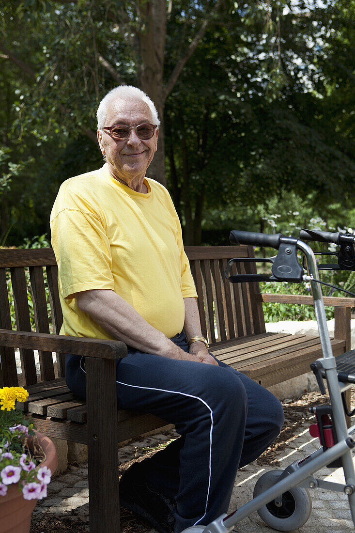 A senior man with a walker resting on a bench in the yard of his nursing home
