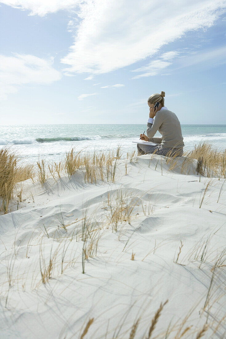 Woman sitting on dunes at beach, using cell phone and taking notes
