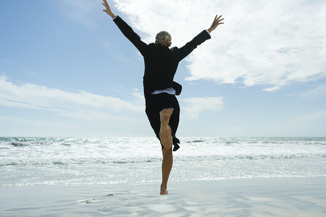 Businesswoman barefoot on beach, jumping into the air, rear view