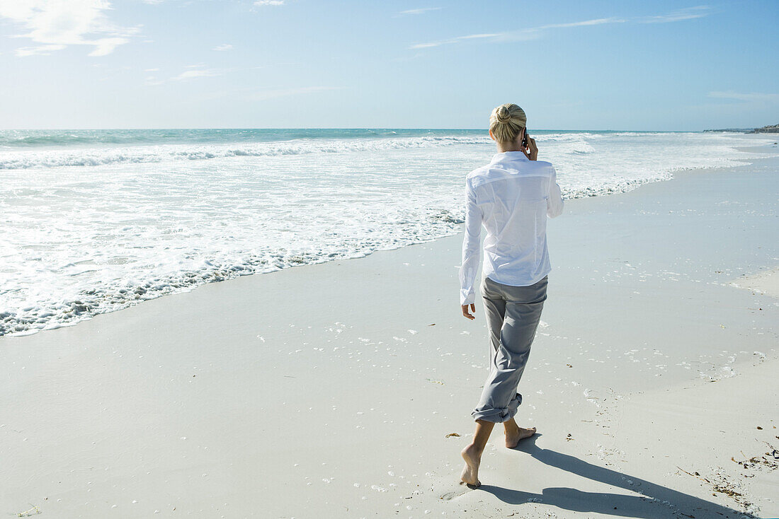 Woman walking barefoot on beach, using cell phone, rear view