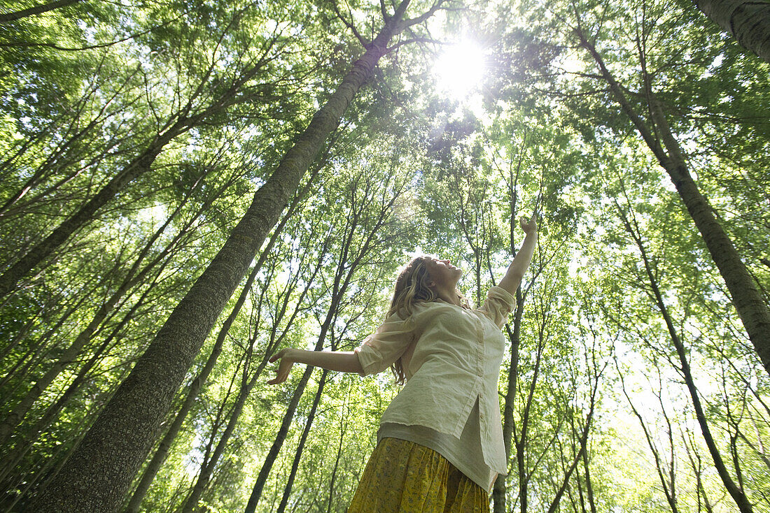 Young woman standing in woods, reaching toward sun shining through canopy