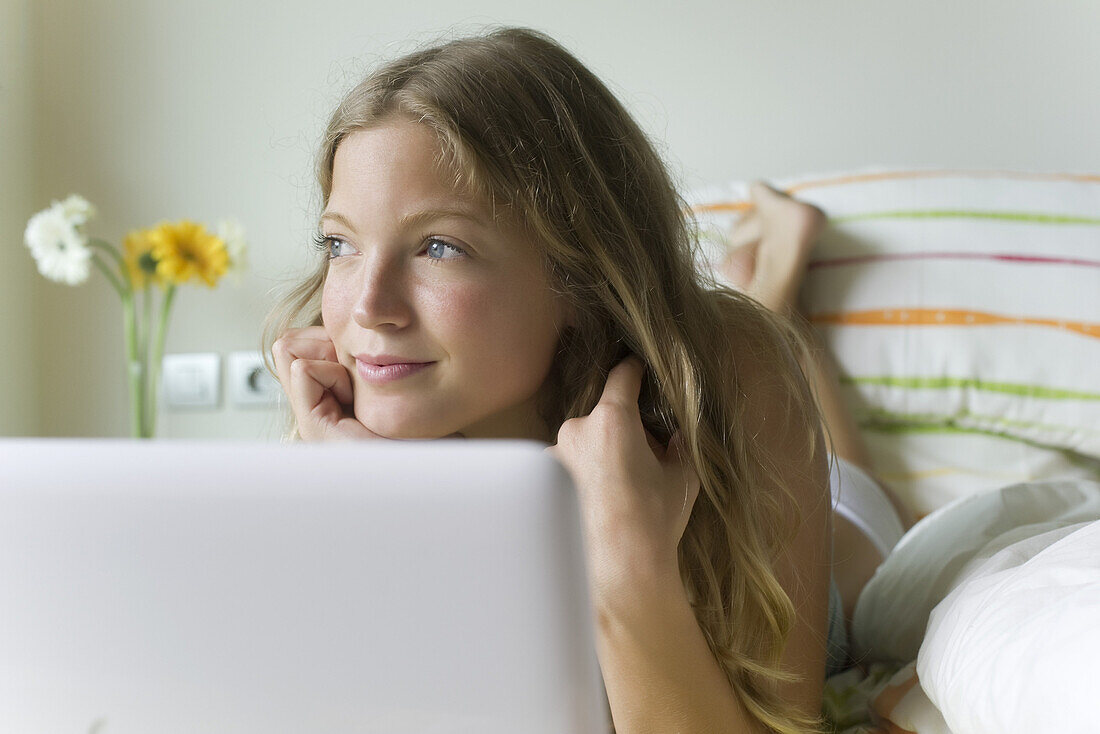 Young woman relaxing in bed with laptop computer, looking away dreamily