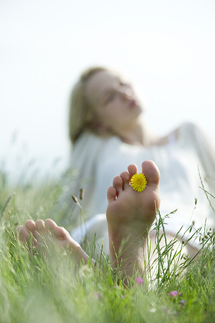 Barfuß im Gras sitzende junge Frau, die eine Löwenzahnblüte zwischen den Zehen hält