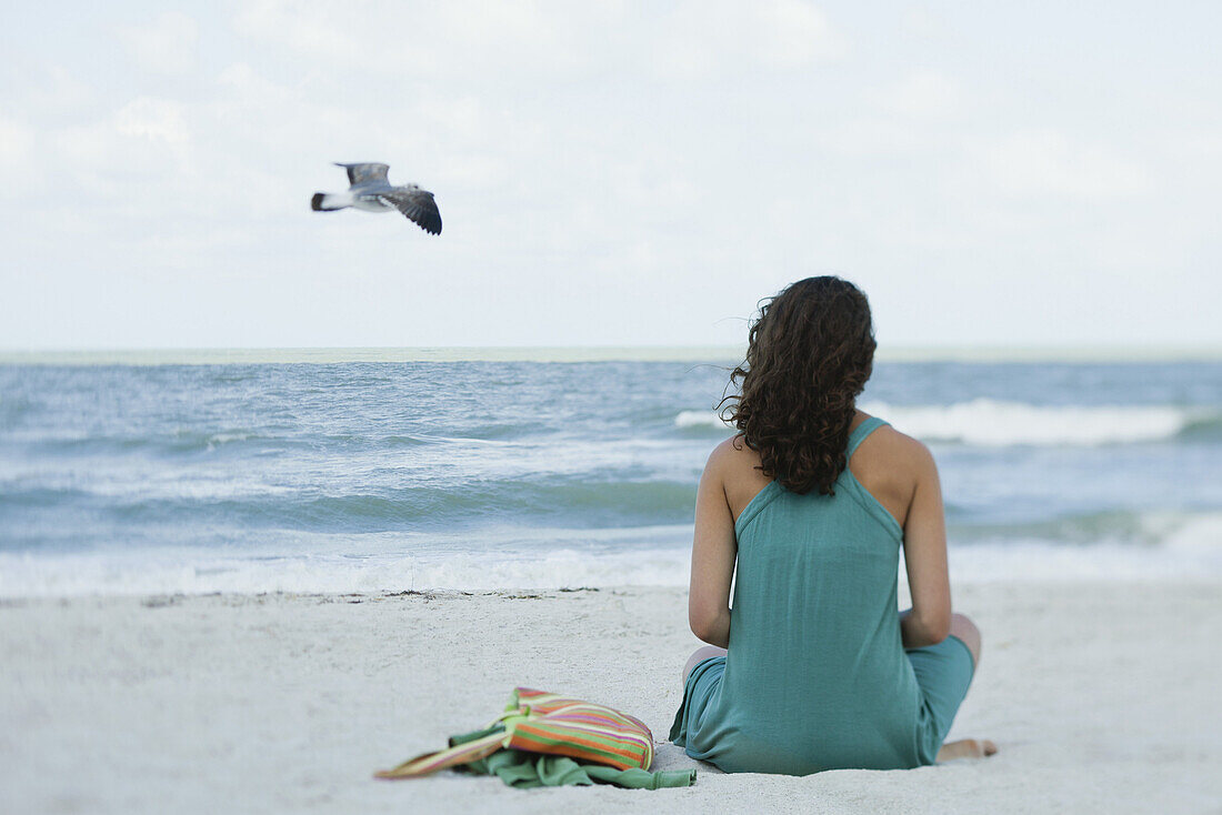 Teenage girl sitting on beach looking at ocean, rear view